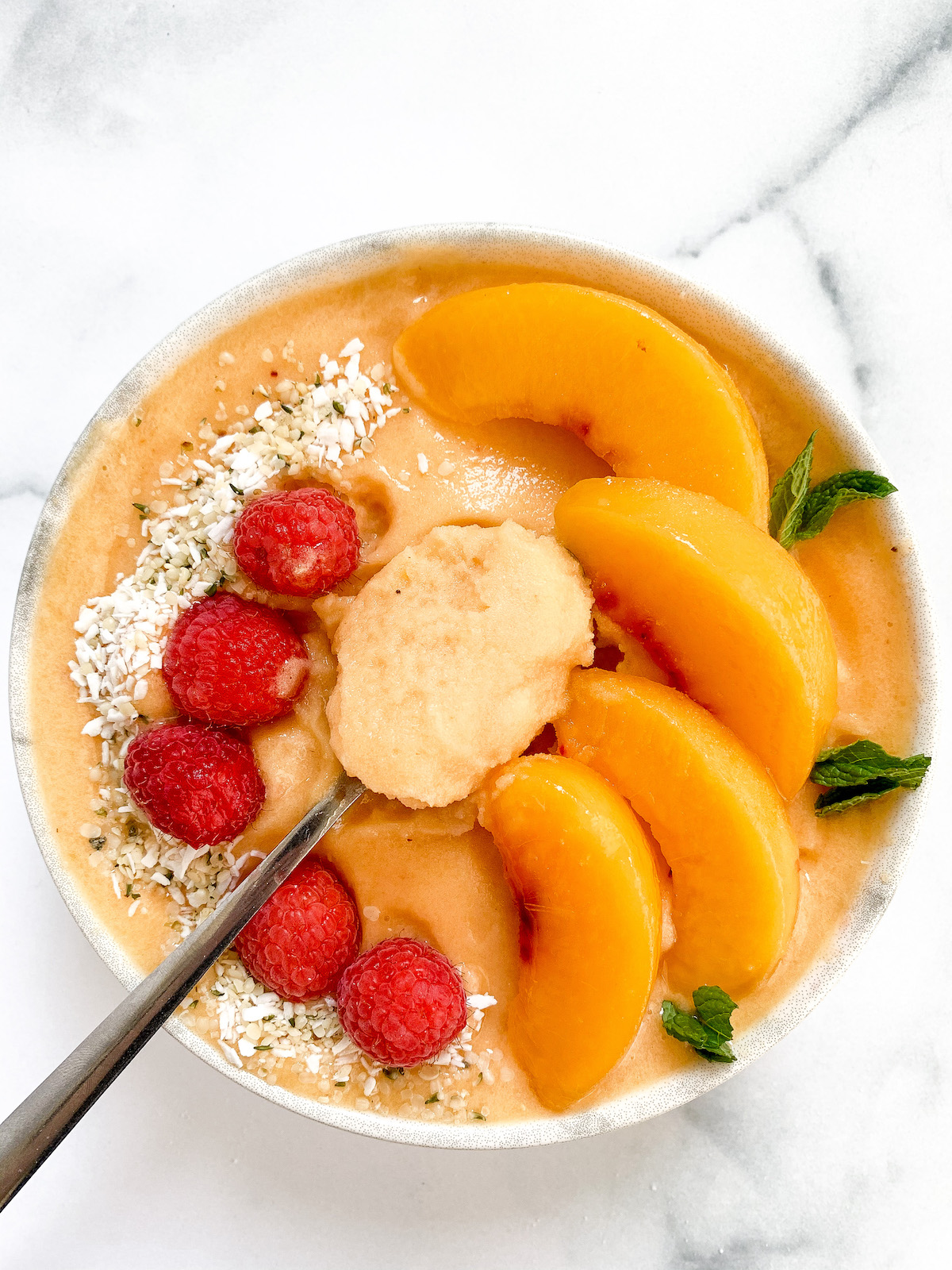 A vegan fuzzy peach smoothie bowl with a spoon topped with peach slices, mint, and raspberries.