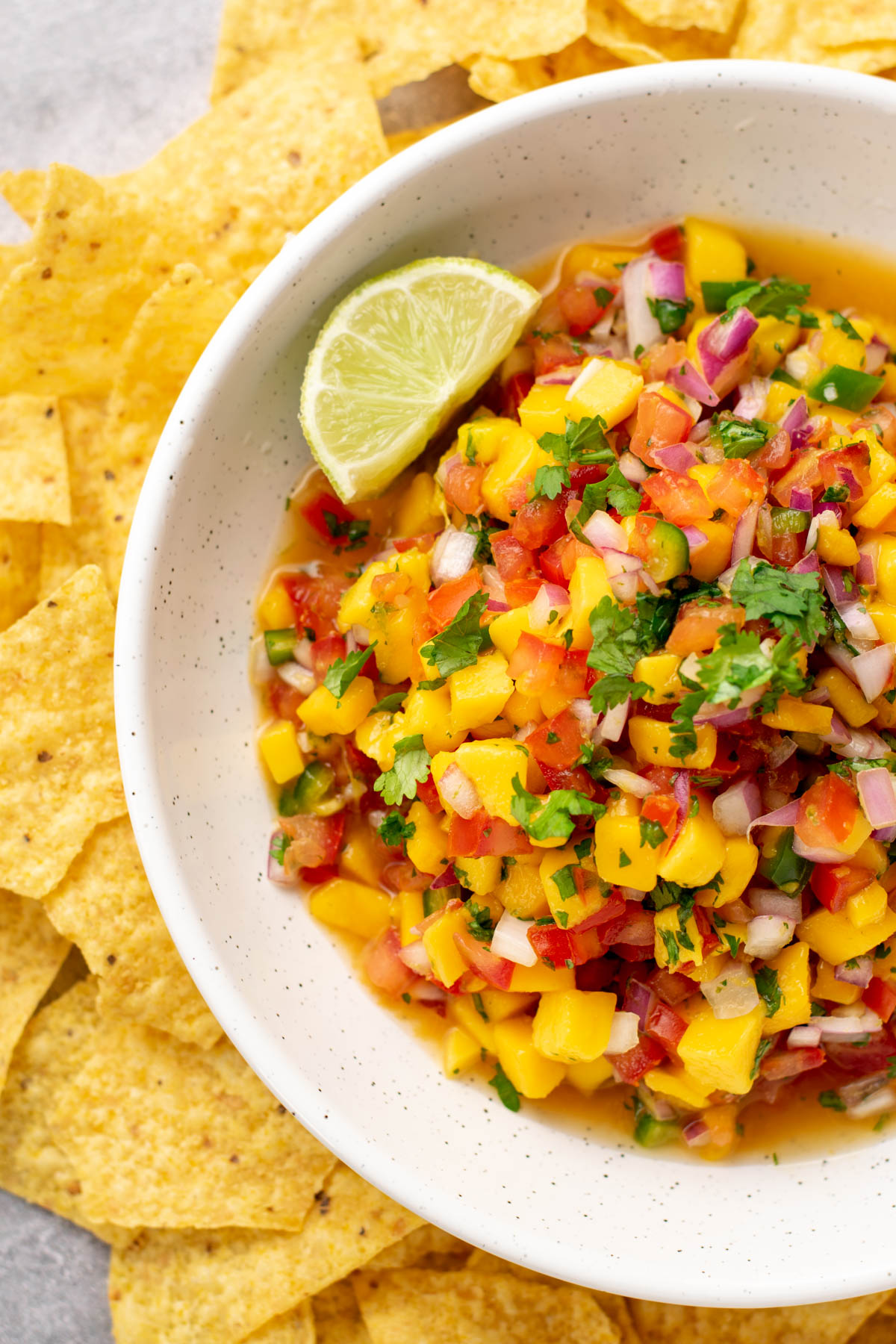 top view of mango pico de gallo in a white bowl with a lime wedge, surrounded by tortilla chips.