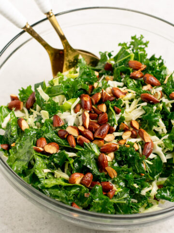 kale salad with almonds on top and gold salad spoons in a glass bowl.